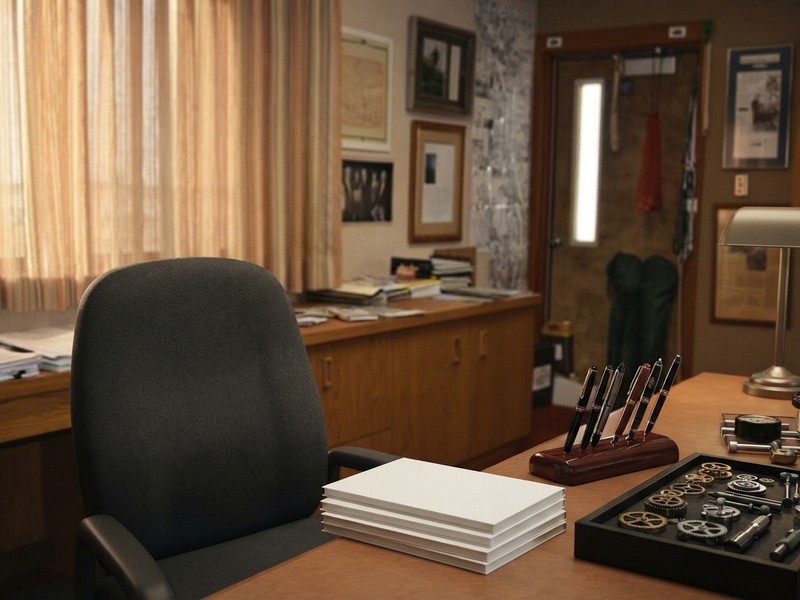 An empty office chair behind a wooden desk with a stack of blank notebooks, a pen stand, and a tray of small mechanical gears.